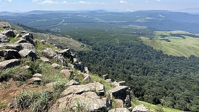 Ein Panoramablick von einem felsigen Hügel auf eine weitläufige Landschaft mit Wäldern, Feldern und sanften Hügeln in der Ferne unter einem strahlend blauen Himmel.